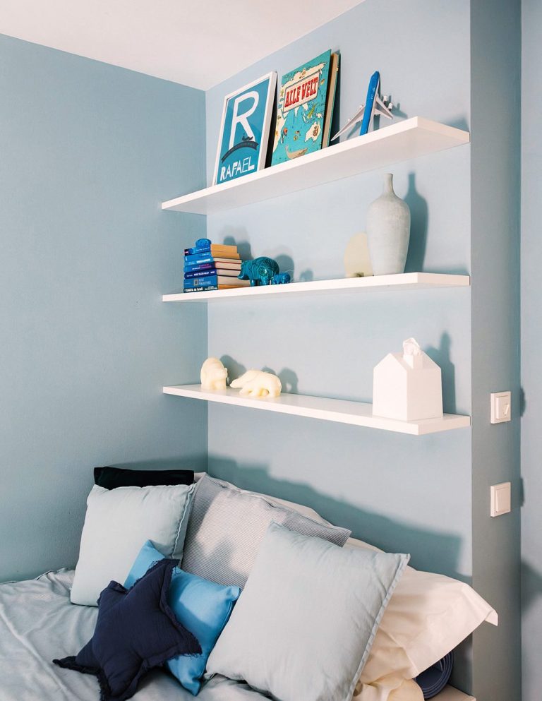 Ein gemütliches Kinderzimmer mit blauen Wänden, maßgefertigten weißen Regalen und blauen Kissen auf dem Bett
